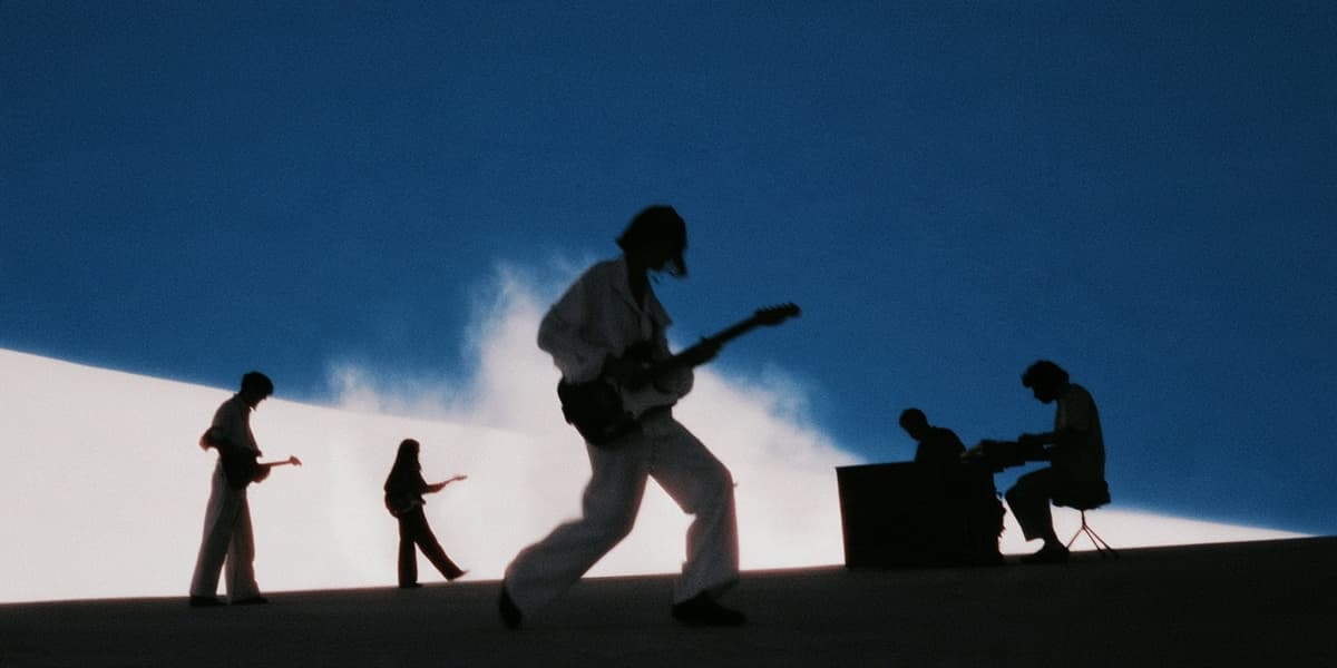 A band in silhouette on a white stage under clouds and blue sky